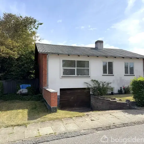 En hvid villa med røde murstensdetaljer står på en grøn græsplæne, omgivet af en hæk og et fladt tag. Der er en garage under huset.