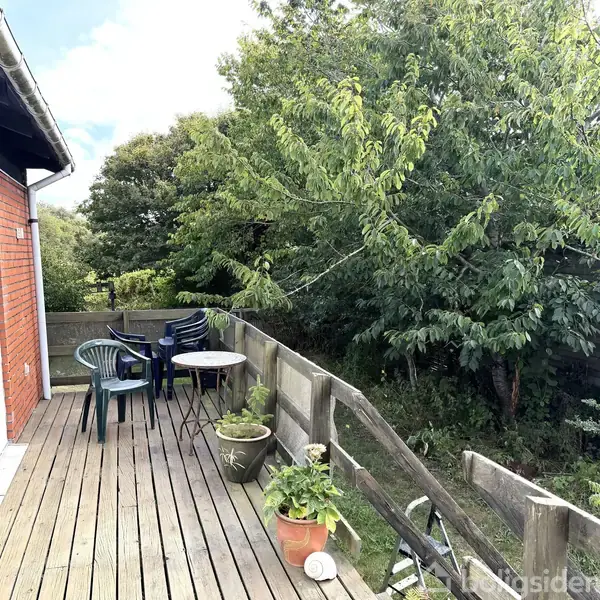 En træterrasse har plasticstole og et lille rundt bord, indrammet af grønne planter og træer i baggrunden, ved siden af et rødt murstenshus.