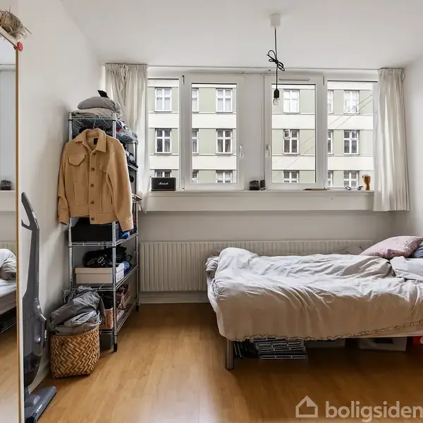 En seng med sengetøj står under tre store vinduer i et lyst værelse. En hylde med tøj og kurve er ved siden af, overfor et spejl.