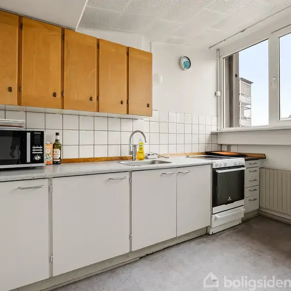 Et køkken med hvidt køkkenskab, træoverflade og backsplash af hvide fliser. Der er en mikrobølgeovn, en flaske olie og opvaskemiddel. Et vindue giver udsigt over byen.