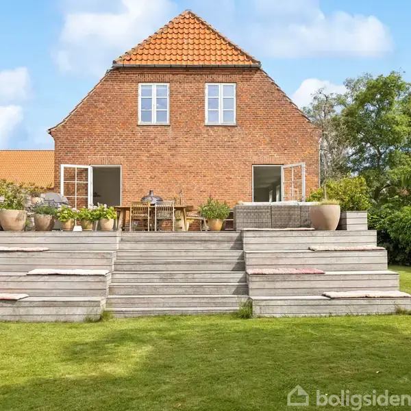 Et murstenshus med rødt tag har en terrasse med potteplanter og udendørs møbler. Trappen fører til en græsplæne, omgivet af træer og en blå himmel.
