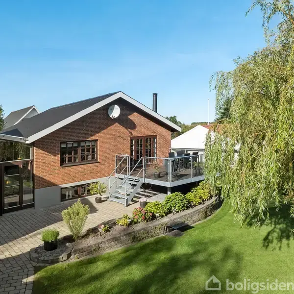 Rødt murstenshus med glasdør og vinduer, trætrappe til hævet terrasse. Omringet af grøn have med græsplæne, buske og et større træ. Blå himmel i baggrunden.