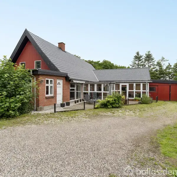 Et rødt murstenshus med gråt tag står stille omgivet af en grusbelagt gårdsplads og grønne buske. I baggrunden ses træer og en blå himmel.