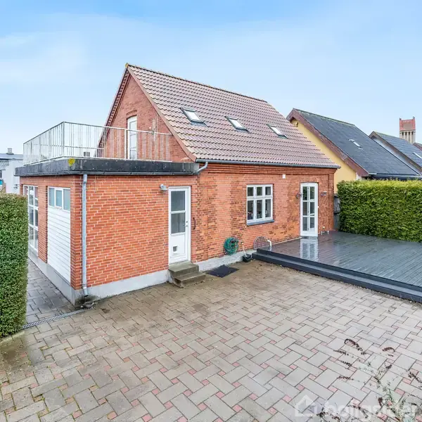 Rødt murstenshus med skråt tag og tagvinduer. Huset står i en flisebelagt indkørsel. Til højre er en terrasse af træ og grønne buske omgiver området.