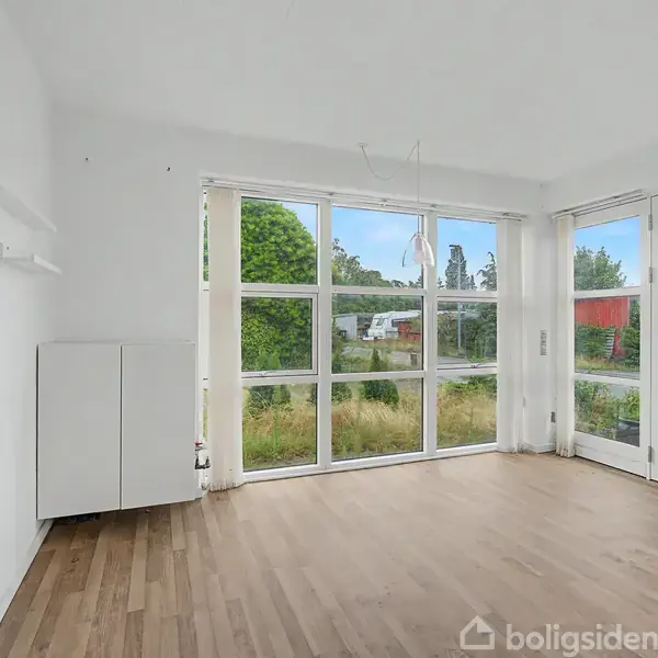 Et lyst rum med trægulv og store vinduer, der lader naturligt lys strømme ind. Rummet har hvide vægge og en dør til højre, med udsigt til grønne omgivelser.