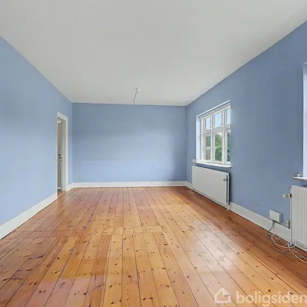 Åben dør leder til en terrasse med betonoverflade og hvidt metalrækværk. Omgivet af grønne træer og med udsigt til et boligområde i baggrunden.