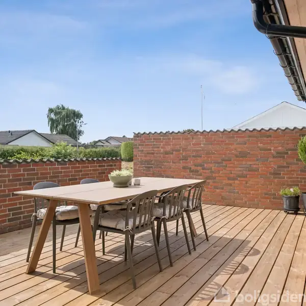Terrasse med træbord og seks stole står på en træbelagt overflade, omgivet af røde murstensvægge. En potteplante er placeret i hjørnet. Klar himmel i baggrunden.