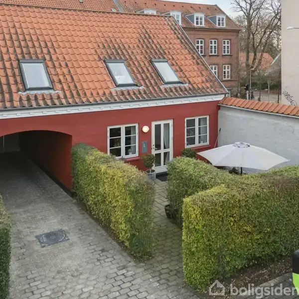 Et rødt hus med rødt tegltag og vinduer. En brostensbelagt indkørsel leder ind under en bue. En hæk omkranser en gårdhave med parasol.