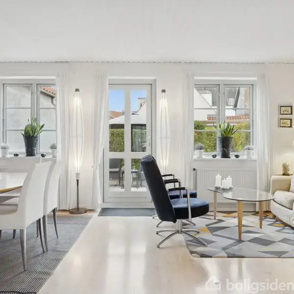 Stue med moderne møblering, inklusiv spisebord med hvide stole, sort lænestol og en lys sofa. Store vinduer og glasdør mod en have, træloftslampe ved sofabordet.