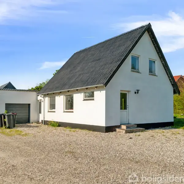 Hvidt hus med sort tag står på en grusvej. Ved siden af ses en garage. Omkring er der grønne buske og træer under en blå himmel.