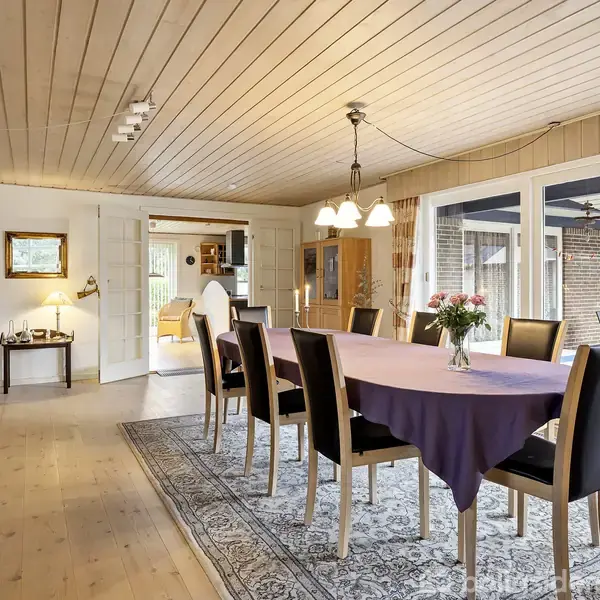 Et spisebord med lilla dug er omgivet af otte stole. En vase med blomster står på bordet. Store vinduer i baggrunden lader lys ind fra terrassen.