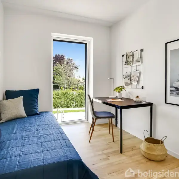 Seng med blå sengetæppe står mod væggen. Skrivebord med stol og lampe står ved vinduet, der viser udsigt til have. Kurv på gulvet kompletterer rummet.