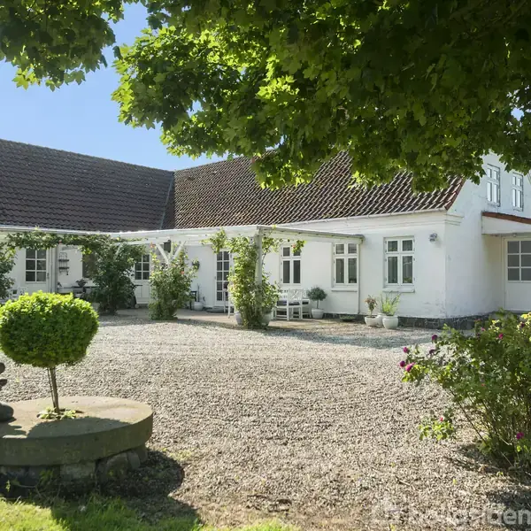 En hvid gård med rødt tag står stille blandt grønne planter og en grusbelagt gårdsplads. Et formklippet træ og en skulptur pynter nær indgangen under en blå himmel.