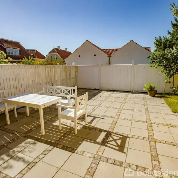 Et hvidt havebord med stole står på en flisebelagt terrasse. Omgivet af et hvidt stakit og en gul mur, med huse i baggrunden under en klar blå himmel.