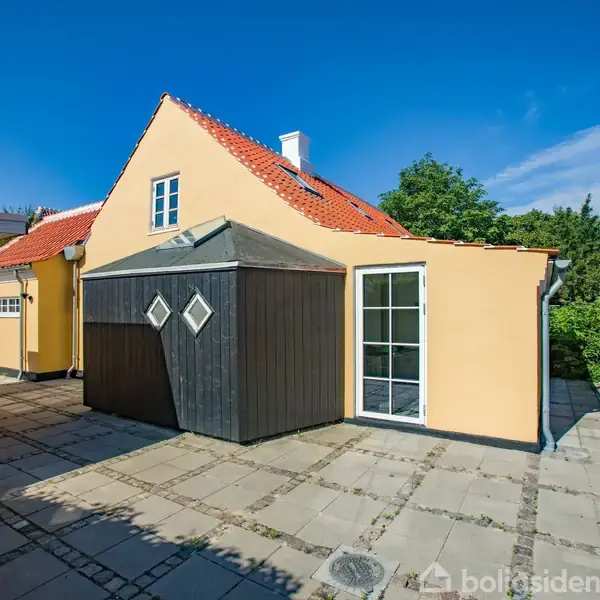 Gult hus med rødt tegltag står stille på flisebelagt gårdsplads. Omgivet af grønt buskads og blå himmel. En glasdør og sort træbeklædt skur er synlige.