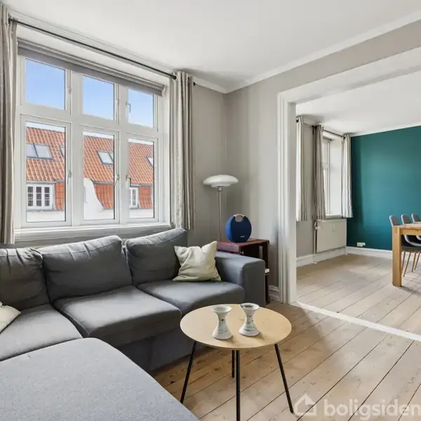 Grå sofa står i stue ved vindue, vindueslys oplyser rummet. Stuen forbinder til spisestue med træbord og stole, grøn væg og kunst maleri i baggrunden.