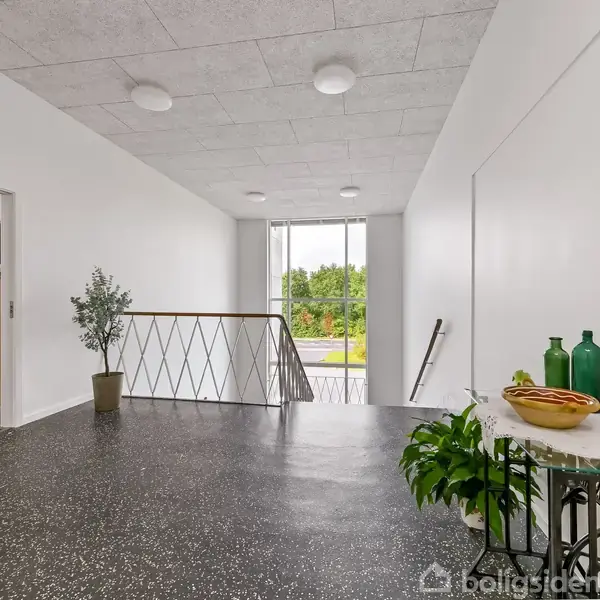 En lys gang med sort marmorlignende gulv, indeholder en plante i en potte samt et bord med dekorative genstande. Et vindue for enden giver udsyn til grønne træer udenfor.