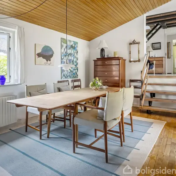 Et spisebord med stole står i et lyst rum med trægulv og hvidmalede vægge. I baggrunden er en trappe til en øvre etage, omgivet af møbler og dekorationer.
