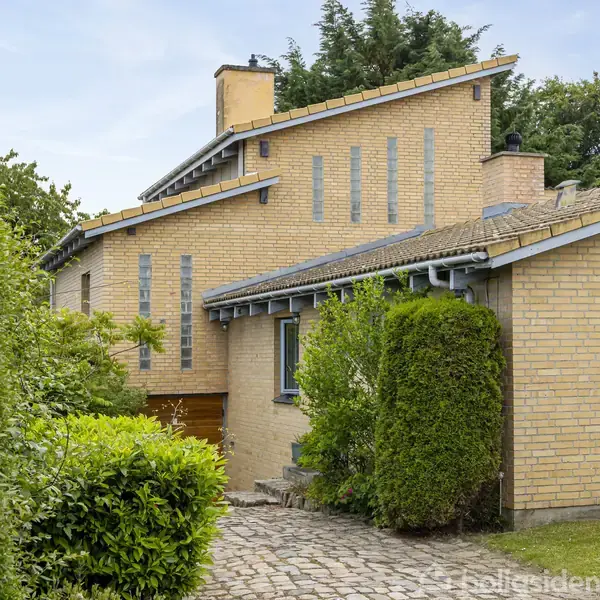 Murstenshus med skrånende tag, omgivet af frodige grønne planter og buske. Huset har flere små vinduer og en velplejet have i forgrunden.