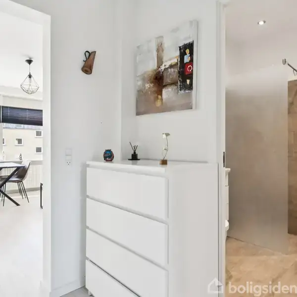 En lys gang med en hvid kommode, udsmykket med kunst på væggen, fører til en moderne spisestue med store vinduer og et stilfuldt badeværelse med fliser.