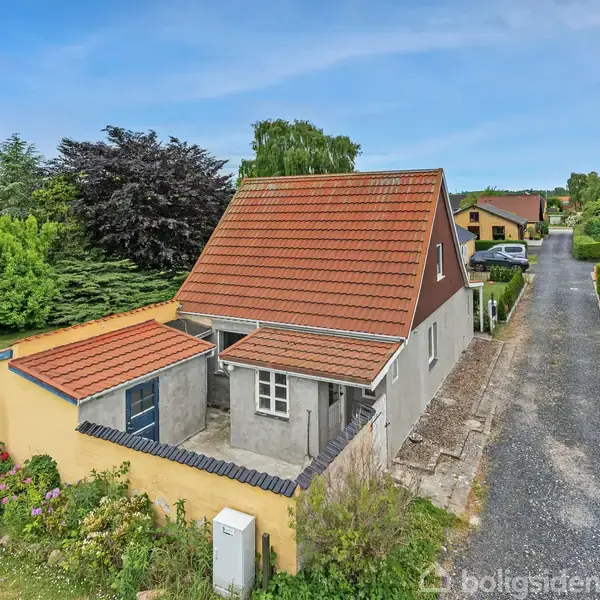 Et hus med rødt tag står i en have. Foran huset er en græsplæne med buske, træer og en terrasse med havemøbler. Himlen er blå i baggrunden.