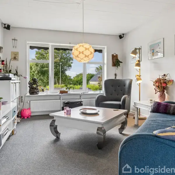 Stue med blåt sofaarrangement, kaffebord og lænestol. En stor vindue giver udsigt til en grøn have. Hylder, lamper og planter udgør resten af rummet.