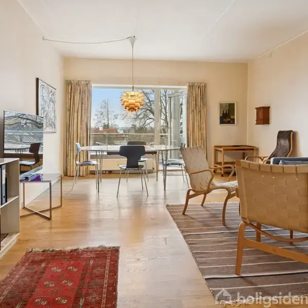 Stue med sofa og to lænestole på tæppe, vender mod spisebord omgivet af stole. Store vinduer og hængelampe giver lys. Reol med bøger og pynt mod venstre væg.