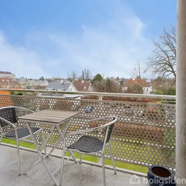 Et sæt af to stole og et bord står stille på en altan med udsigt til en bydel med huse og træer under en klar blå himmel.