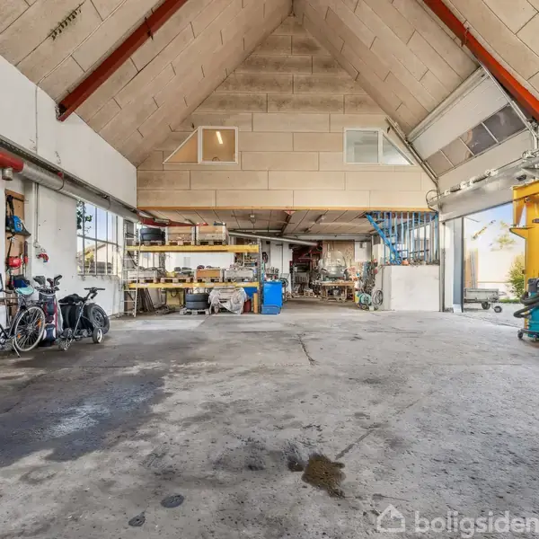 En rummelig værkstedshal med højt loft og store vinduer. Værkstedet har cykler og diverse værktøjer langs væggene. Gennem gulvet ses nogle pletter. To-etagers struktur mod bagvæggen.