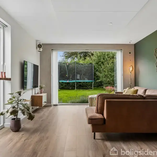 En brun sofa står i en stue med trægulv, ved siden af en vægdekoration. En stor åbning viser en have med en trampolin. Lysindfald gennem høje vinduer.
