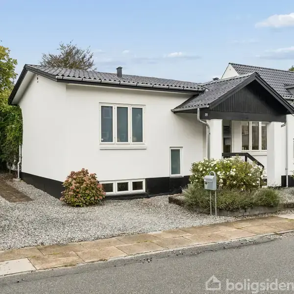 En hvid enetages villa med mørkegråt tag. Forhaven er grusbelagt, omgivet af buske, og der er en indkørsel til venstre. Huset står på en stille gade.