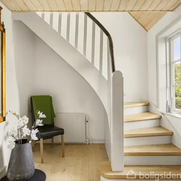 Trappe fører opad i et lyst rum med træloft, træpaneler og stort vindue. Ved siden af trappen står en stol og en dekorativ vase med blomster.irection