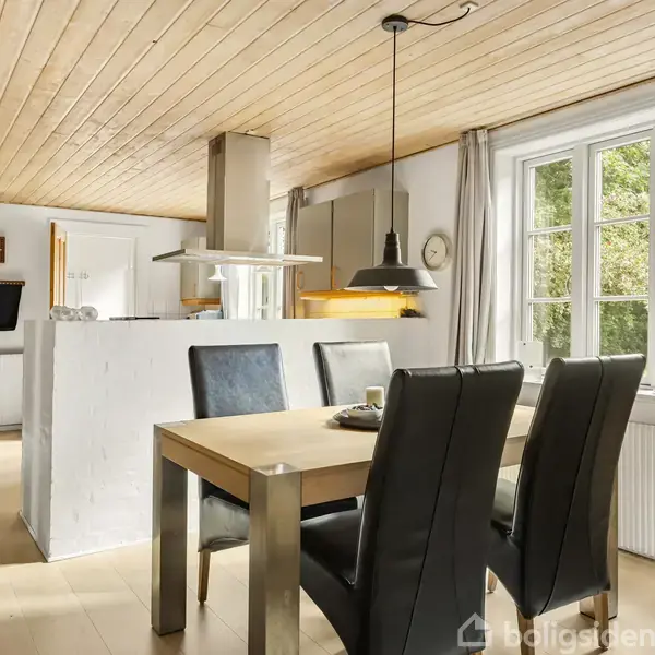 Et træbord med fire sorte lænestole står i et lyst køkken-alrum med trægulv og -loft. Et stort vindue med gardiner giver udsigt til grønne omgivelser.