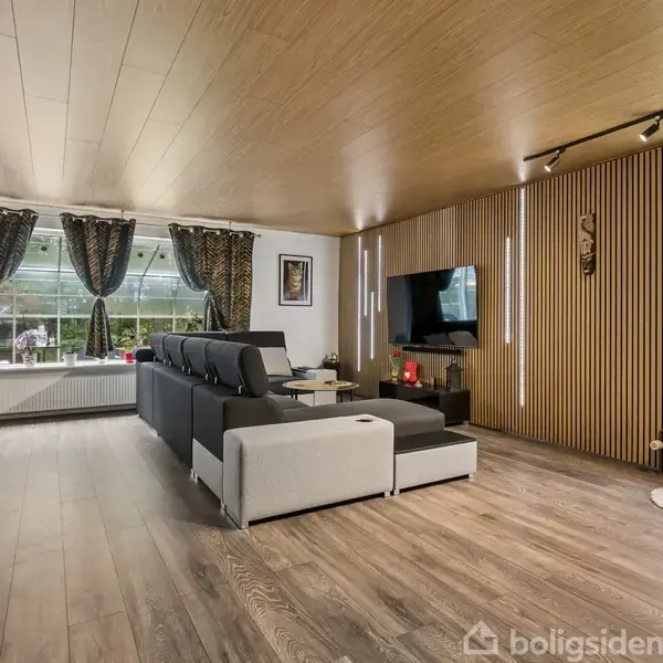 En stor, moderne stue med en grå sofa centreret mod en vægmonteret tv-skærm. Rummet har et træloft og gulv, med store vinduer dækket af tunge gardiner, der tillader naturligt lys.