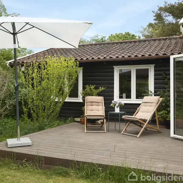 En terrasse med to liggestole under en stor parasol. Ved siden af er en sortmalet træbygning med hvide vinduer, omgivet af grøn beplantning.