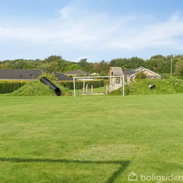 En legeplads med en bakke, tunnelrør og klatrestativer omgivet af velplejet græsplæne. I baggrunden ses huse og træer under en blå himmel.