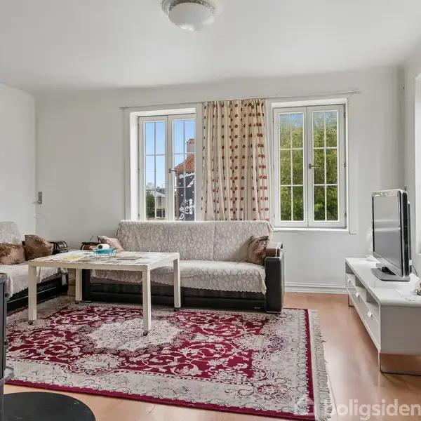En stue med en sofa og sofabord, hvor en fladskærms-tv står på en skænk. Rummet er lyst med store vinduer, og en rød mønstret tæppe dækker gulvet.