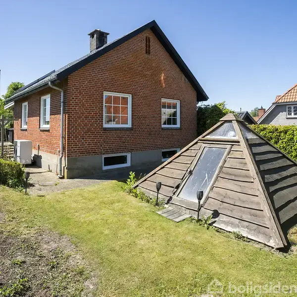 Rødt murstenshus står i en grøn have, omgivet af hække. Ved siden af huset er en pyramideformet trækonstruktion med glas. Huset har hvide vinduer og sort tag, og himlen er klar.
