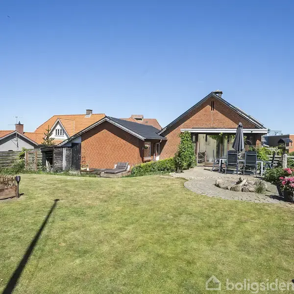 En gulgrøn græsplæne omgiver et rødt murstenshus med udendørs terrasse. Hvide stole og et bord står på terrassen, flankeret af blomster og buske. Klar blå himmel.