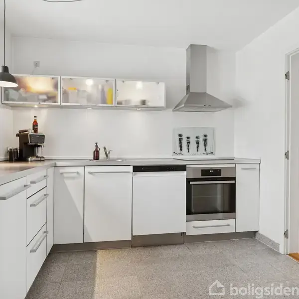 Et moderne køkken med hvide skabe arrangeret i en L-form. Over bordpladen hænger en lampe, og der er et vindue til venstre. En ovn og emhætte er midtpunktet.