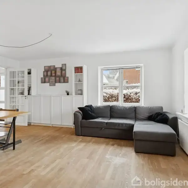 Stue med grå sofa ved siden af tre vinduer, spisebord med stole under en hængende lampe, maleri og reol på væggen, trægulv, lys atmosfære.