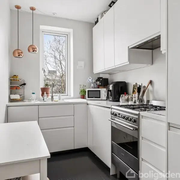 Et hvidt køkken med bordplade og skabe langs væggen. En ovn og mikroovn er placeret ved siden af hinanden. Et vindue giver naturligt lys.
