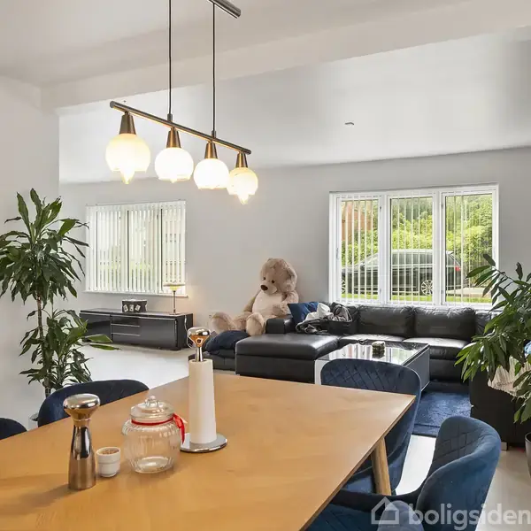 Et spisebord med stole står i en lys stue med planter. En stor teddybjørn sidder på en sort sofa ved vinduer med udsigt til grønt udenfor.