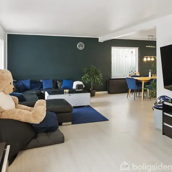 Stor bamse sidder på sort sofa i en moderne stue med TV, grønne planter og spisebord i baggrunden. Rummet har et lyst trægulv og mørkeblå væg.