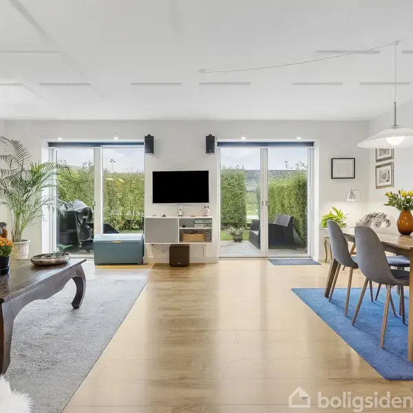 Stue med et spisebord til højre, en sofagruppe til venstre, og et TV i midten. Store vinduer åbner ud til en terrasse med grøn beplantning udenfor. Ingen tekst.