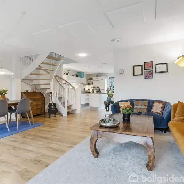 En stue med trægulv, hvor en blå sofa og en sennepsfarvet sofa står omkring et træbord. En spiseplads med bord og stole står ved trapper, der fører opad. En lampe og vægbilleder fuldender rummets indretning.