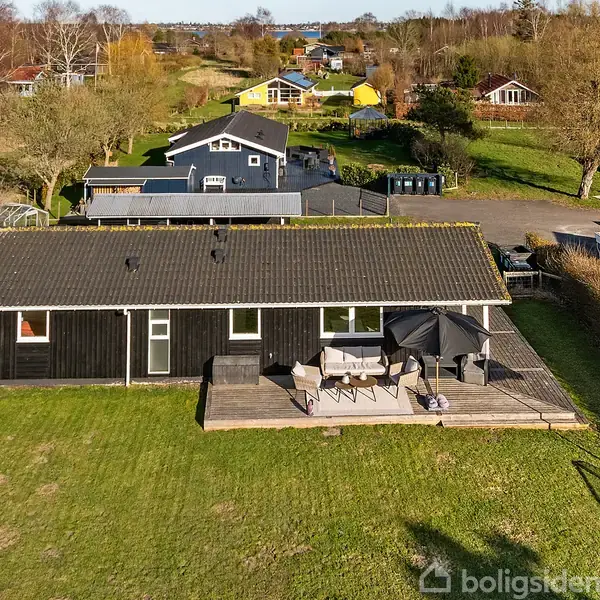 Et sortmalet sommerhus med en terrasse, hvor der står borde og stole. Placeret i en grøn have med en dansk flagstander. Omgivet af andre huse og træer.