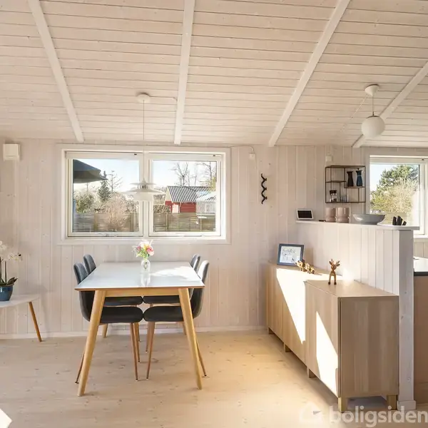 Spisebord med fire stole står foran et vindue. Bordet har en vase med blomster. Køkken med lyse skabe og grå bordplader er til højre i et lyst, skandinavisk rum.