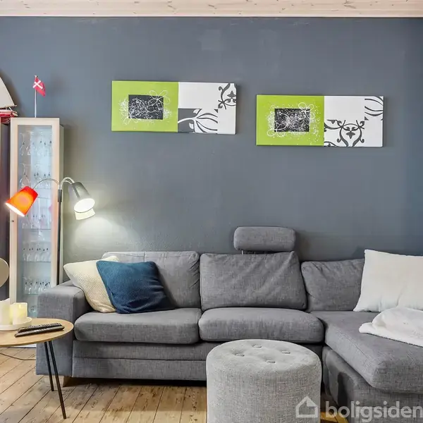 Grå hjørnesofa står i en stue mod en mørkeblå væg, med to malerier ovenfor; til venstre står et vitrineskab og en lille træbordlampe. Der er ingen tekst.