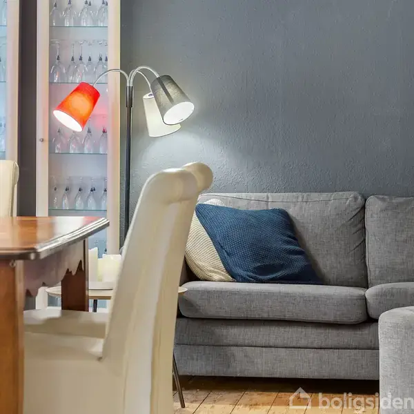 Grå sofa med blå pude står op ad en mørk væg. Ved siden af er der en gulvlampe med rød og hvid skærm. Trægulv og spisebord til venstre.